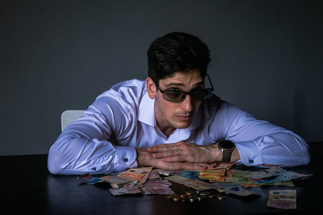 Portrait of a man in sunglasses leaning over a table covered with various currencies.
