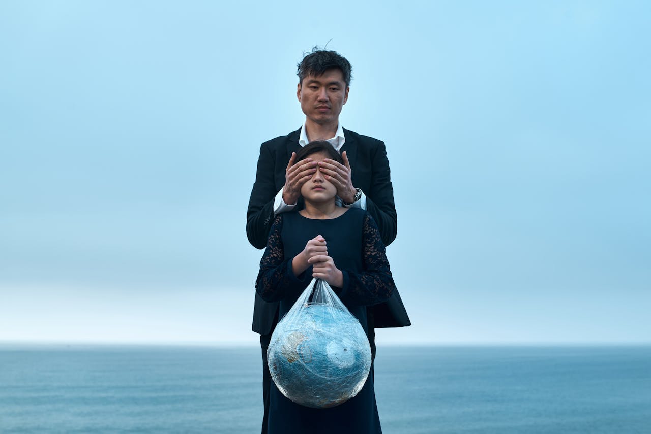 our-journey-02 Asian male in formal wear standing near seashore and covering eyes of girl in black dress with Earth globe in plastic bag environment pollution concept