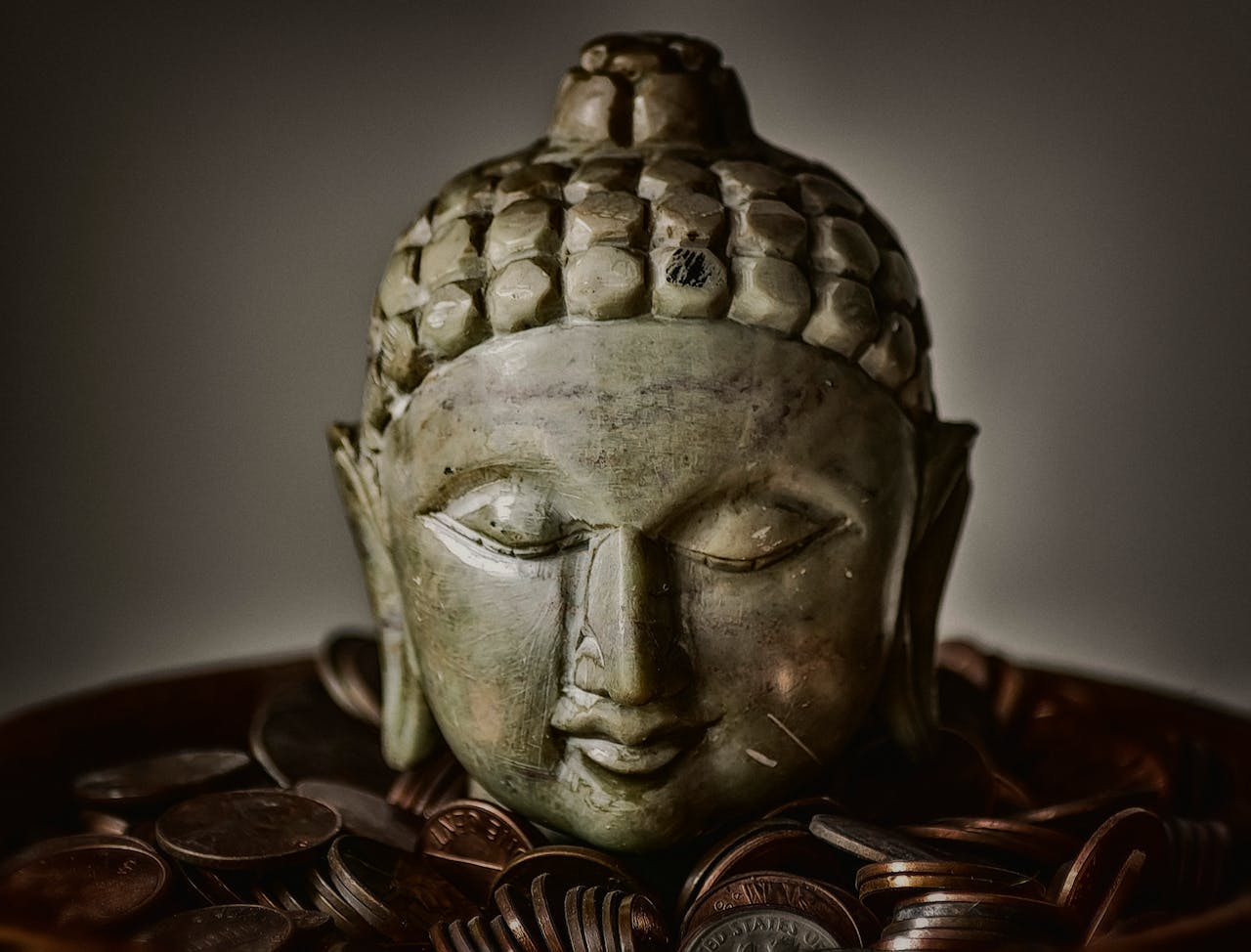 Close-up of Buddha head sculpture resting on bronze coins, symbolizing spirituality and prosperity.